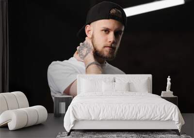 Stylish bearded caucasian young man with tattooed arm in cap and white t shirt looking at camera, light smiling, touching neck with hand on dark background with electric diode lamps. Fashion concept Wall mural