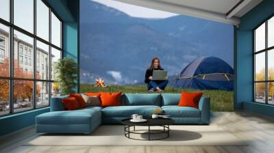 young female freelancer working on laptop in the mountains in the evening. Tourist girl sitting near campfire and tent. Copy space Wall mural