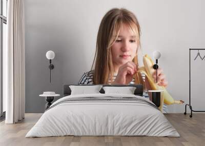 A little hungry girl eats a ripe yellow banana on a white background. The girl bites a banana Wall mural