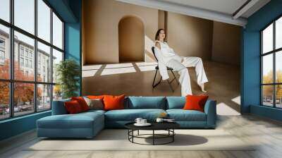 Front view of confident female model ,dressed in white formal suit and heels, sitting on chair and posing at camera on background of wall with minimalism design elements Wall mural