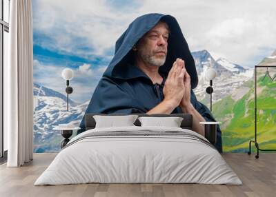 An elderly monk in a black hooded cassock meditates and prays among alpine mountains under a blue sky Wall mural