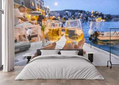 Raising hands holding a glass of white wine in front of a seaside village at a greek restaurant. Wall mural