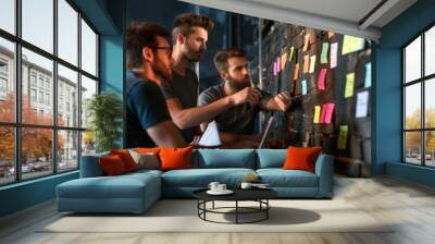 A group of three men collaborates in a coworking space, discussing ideas while reviewing colorful sticky notes attached to a brick wall. A laptop and notebooks are present on the table Wall mural