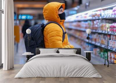 A person dressed in an orange puffer jacket and wearing a black mask is standing in a supermarket aisle, carefully examining products on the shelves while carrying a backpack Wall mural