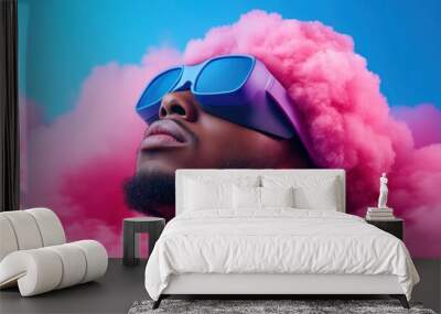 A young man gazes upwards, showcasing his bold pink cloud-like hairstyle alongside trendy sunglasses. The striking blue background enhances the vibrant atmosphere of artistic expression Wall mural