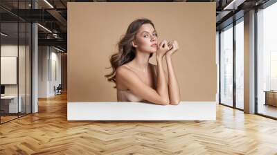 Beautiful blonde curly hair in a beige bodysuit is sitting at a white table. Perfect slim body natural makeup. Long hair, natural cosmetics Wall mural