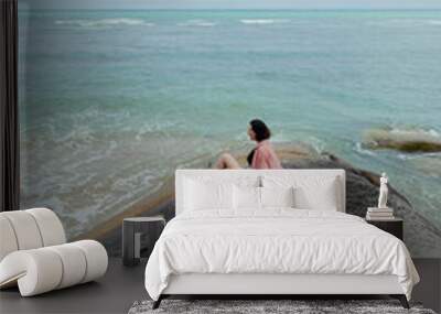 Solitary woman rests on a rock by the sea, looking out at the tranquil waters Wall mural