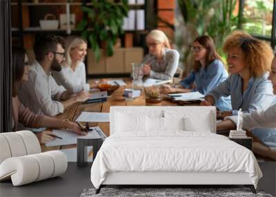 Team members gather around a wooden table to exchange ideas and discuss strategies. The atmosphere is collaborative, with notes and drinks scattered across the table Wall mural