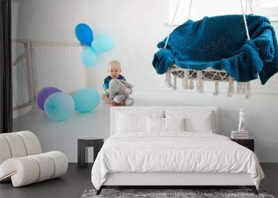 a little boy sits on the floor with a soft toy among the balloons and near the hanging chair Wall mural