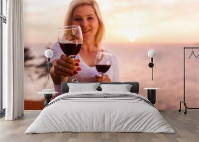 Drinking together. Low angle view of beautiful young couple clinking glasses with wine while sitting on the beach with sunlight on background. Wall mural