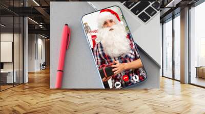 holidays, technology and people concept - man in costume of santa claus in smartphone, presents and christmas tree sitting in armchair over lights background Wall mural