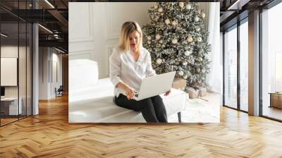 Young pretty blond woman shopping online using white laptop against Christmas background. Wall mural