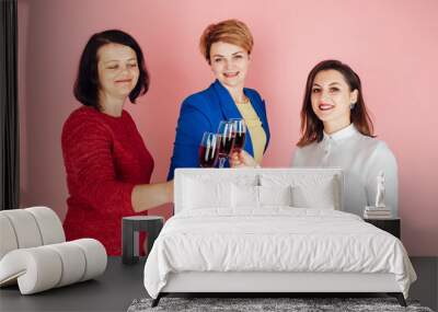 Studio shot of three women drinking alcohol. Beautiful women with a glass of wine in their hands. Pink background. Maiden evening. Women's Day. Girlfriends celebrate Wall mural