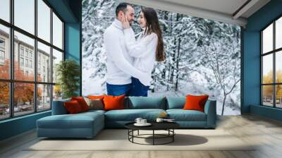 winter love story. snow-covered pine forest. man and woman hugging in the winter forest. girl on a guy dressed in a white winter sweater in a white snowy forest Wall mural