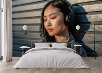 A young Asian woman wearing headphones listens to music with her eyes closed, standing on the street near the wall of the house. Wall mural