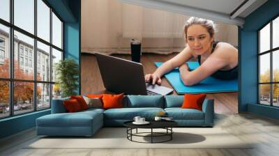 A sporty woman in sportswear is sitting on the floor with dumbbells and a protein shake or a bottle of water and is using a laptop at home in the living room. Sport and recreation concept. Wall mural