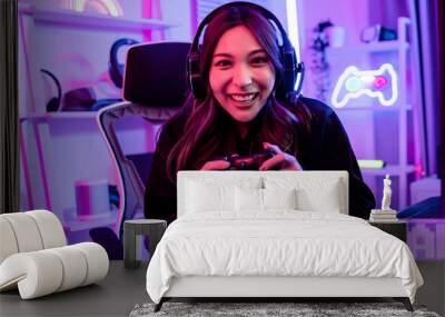 Beautiful happy young female game online streamer or caster smiling with fun, succeed for winner, using joinstick to play or compete, sitting in room with decorated neon light. Looking at camera. Wall mural