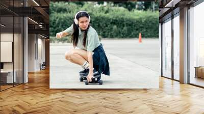 Beautiful sportive hipster happy Asian woman wearing casual shirt and shorts with headphone to listen music while playing skateboard as hobby in free time day on holiday, smiling at outdoor park. Wall mural