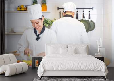 Two Asian professional couple chef wearing white uniform, hat, helping each other, preparing meal,cooking in kitchen together, smiling with happiness and confidence. Food Concept. Wall mural