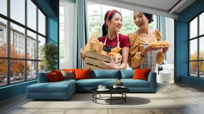 Two beautiful Asian women smiling with happiness and confidence, holding and presenting wooden container of basket of bread, croissant, baguette for bakery in indoor kitchen at home. Hobby Concept. Wall mural