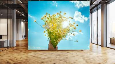 A bouquet of field daisies in hand against the background of the sky and sun. Wall mural