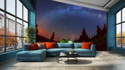 Night photo of the sky and the Milky Way. Wildlife, in a clearing in the mountains there are two glowing tent among the tall silhouettes of dark firs
 Wall mural