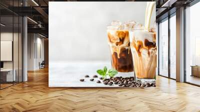   A pitcher pours coffee into two glasses filled with ice and coffee beans on a table with green leaves Wall mural