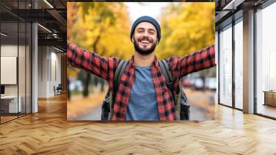 A cheerful man stands in a city park adorned with golden autumn leaves, wearing a beanie and a plaid shirt, expressing joy with his arms outstretched in celebration of the beautiful weather Wall mural