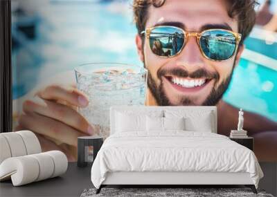 A cheerful young man relaxes poolside, holding up a glass of sparkling water, reflecting the serene blue water and sunny sky in his sunglasses Wall mural