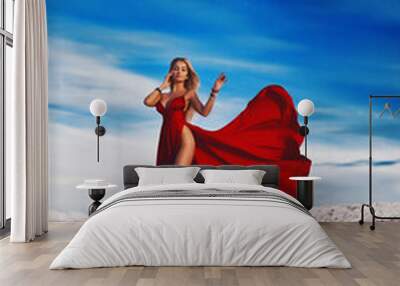 A gorgeous blonde girl in a sexy red dress with deep slits on the hips against the background of a white sandy beach and a sunset sky. Wall mural