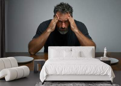 A man in casual attire sits at a wooden table with empty plates surrounding him, displaying an expression of frustration and hunger in a subdued indoor setting Wall mural