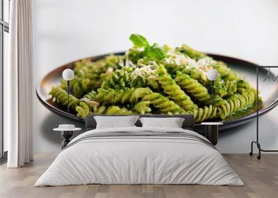 A plate of fusilli pasta covered in green pesto sauce, garnished with grated Parmesan cheese and a sprig of basil, served on a dark-colored plate against a light background. Wall mural