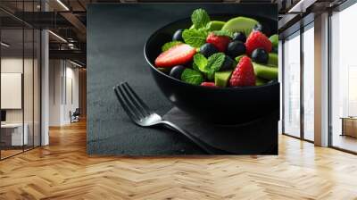 A vibrant fruit salad made with fresh strawberries, blueberries, kiwi slices, and mint leaves is presented in a black bowl. The dark background enhances the salad's colorful appearance Wall mural