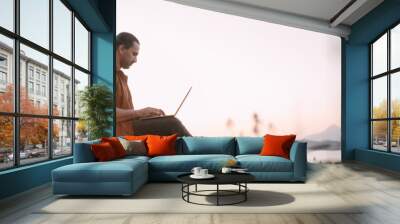 A young Caucasian man remotely works with a laptop in a garden on a mountain overlooking the sea and sunset. Wall mural