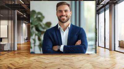 A young man with a well-groomed beard stands confidently with arms crossed, wearing a blue suit and a white shirt, smiling in a bright, contemporary office with greenery around Wall mural