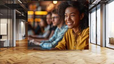 A young woman with curly hair smiles while using her laptop in a lively cafГ©. The ambient lighting creates a warm atmosphere as other students concentrate on their studies nearby Wall mural