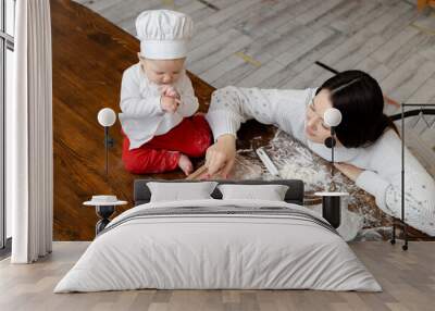 An Asian baby in a chef 's hat and apron with a young mother cook Christmas ginger cookies at home on the kitchen table and smile happily Wall mural