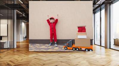 cute toddler boy in a red overalls and a cap with a bright emotion of joy near a large toy car - a truck with many cardboard boxes. Express - parcel delivery, postman, truck driver. Successful deal Wall mural