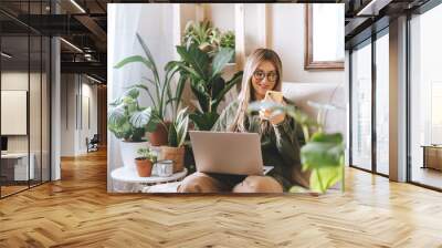 Freelance woman in glasses with mobile phone typing at laptop and working from home office. Happy girl sitting on couch in living room with plants. Distance learning online education and work. Wall mural