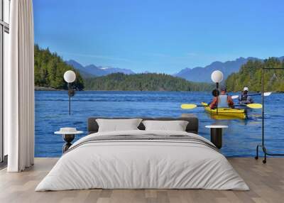 Group of friends on sea kayak in Pacific Ocean near Vancouver Island. Colorful kayaks, trees on the island, man with west, hat and paddle. Blue sky.  Wall mural