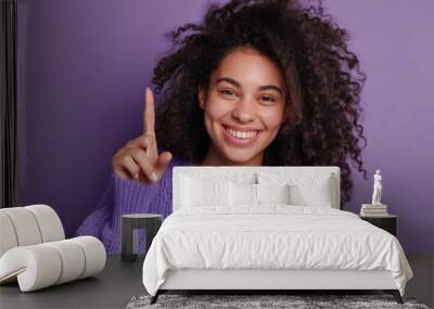 Happy smiling woman with curly hair wearing purple sweater pointing at something with finger Wall mural