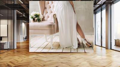 legs of the bride in white shoes and a dress that stands near the pink chair on which lies a wedding bouquet on the background of a gray wall Wall mural