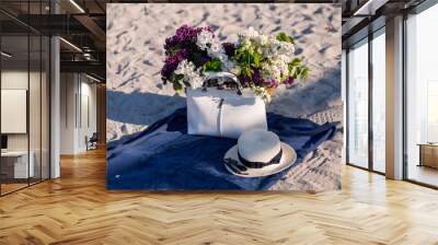 Lilac bouquet on the summes beach. Female straw hat, sunglasses and white stylish bag on a blue cover. Sandy beach background. Wall mural