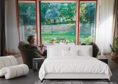 A little girl and her grandfather drinking tea, seated on bean bags, enjoying view of a lush garden through a large glass door Wall mural