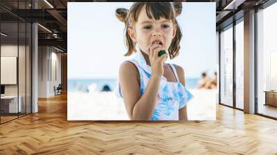 Little girl sitting on the beach and eating a cucumber Wall mural