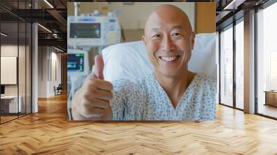 Middle-aged Asian bald patient after chemotherapy in hospital gown giving thumbs up with medical equipment in background Wall mural