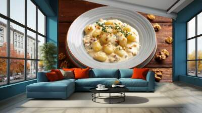 Overhead shot of gnocchi with creamy sauce, topped with chopped walnuts and herbs, served in a white bowl on a rustic wooden surface, with scattered walnuts and fresh thyme. Wall mural