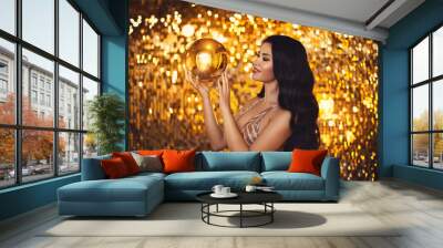 Portrait of a young brunette girl in an evening dress with a golden Christmas ball in her hands on a gold background Wall mural