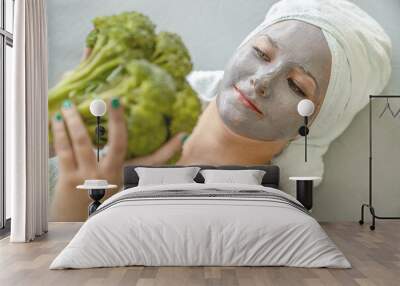 Concept of natural facial skin care. Funny young woman with cosmetic mask made of gray clay, her hair and body wrapped in towels, she holds broccoli in her hands, portrait, close-up. Spa procedure.  Wall mural