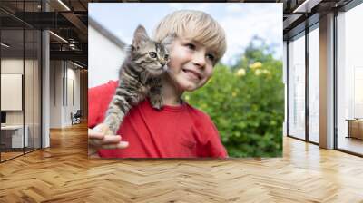 striped fluffy kitten sits on the shoulder of a cute boy of 7 years old, looking in one direction. cat day. happy childhood. Gentle positive atmosphere of communication between animals and people Wall mural